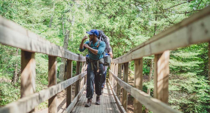 A person flexes their muscle as they walk across a wooden bridge toward the camera. There are dense green trees behind them.